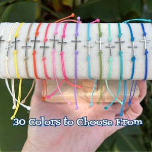 May include: A collection of thin cord bracelets, each featuring a small silver-toned cross charm. The bracelets are displayed in a variety of vibrant colors including yellow, orange, pink, purple, green, and blue. Text reads: 30 Colors to Choose From.