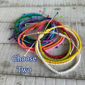 May include: A pile of colorful beaded bracelets in various colors, including white, yellow, red, pink, orange, teal, purple, and blue. The text "Choose Two" is written below the bracelets.