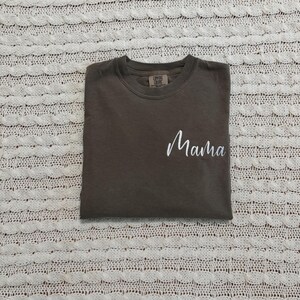 May include: A folded, dark brown t-shirt with the word "Mama" in white script on the front. The shirt has a crew neck and a small tag on the upper back. The shirt is on a white, textured surface.