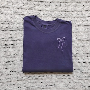 May include: A folded, indigo-colored t-shirt with a crew neck. A light purple bow and the letter "L" are embroidered on the front. The shirt is displayed on a gray, textured surface.