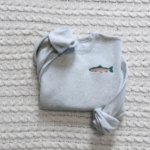 May include: A light gray crewneck sweatshirt featuring a small, detailed embroidered graphic of a trout fish on the left chest. The fish is depicted with orange, green, and black thread colors, showcasing a realistic profile view of the aquatic creature.