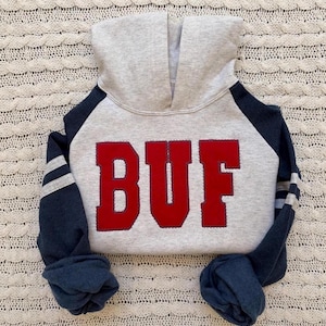 May include: A light gray hoodie with dark navy blue raglan sleeves and a hood. The front features the letters BUF in large, bold red fabric lettering with visible stitching. Each navy blue sleeve has two horizontal light gray stripes near the cuff.