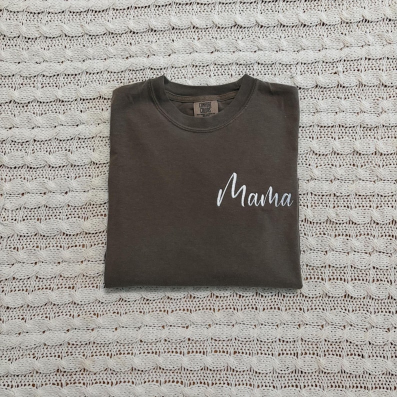 May include: A folded, dark brown t-shirt with the word "Mama" in white script on the front. The shirt has a crew neck and a small tag on the upper back. The shirt is on a white, textured surface.