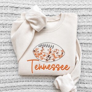 Puede incluir: Una sudadera de cuello redondo color crema con una forma de bal&oacute;n de f&uacute;tbol bordada y rellena de motivos florales naranjas y marrones. Debajo del bal&oacute;n, la palabra Tennessee est&aacute; bordada en letras cursivas naranjas en la parte frontal.