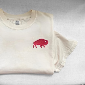 May include: Cream-colored t-shirt with a red buffalo emblem embroidered on the front. The shirt is folded, showing the Comfort Colors label. The short-sleeved tee is made of soft material.