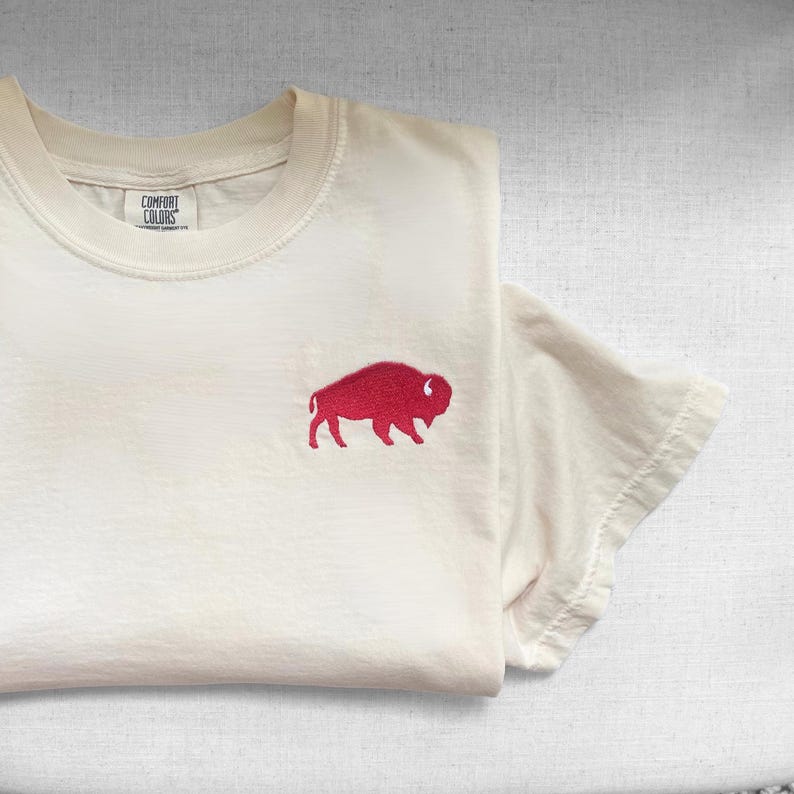 May include: Cream-colored t-shirt with a red buffalo emblem embroidered on the front. The shirt is folded, showing the Comfort Colors label. The short-sleeved tee is made of soft material.