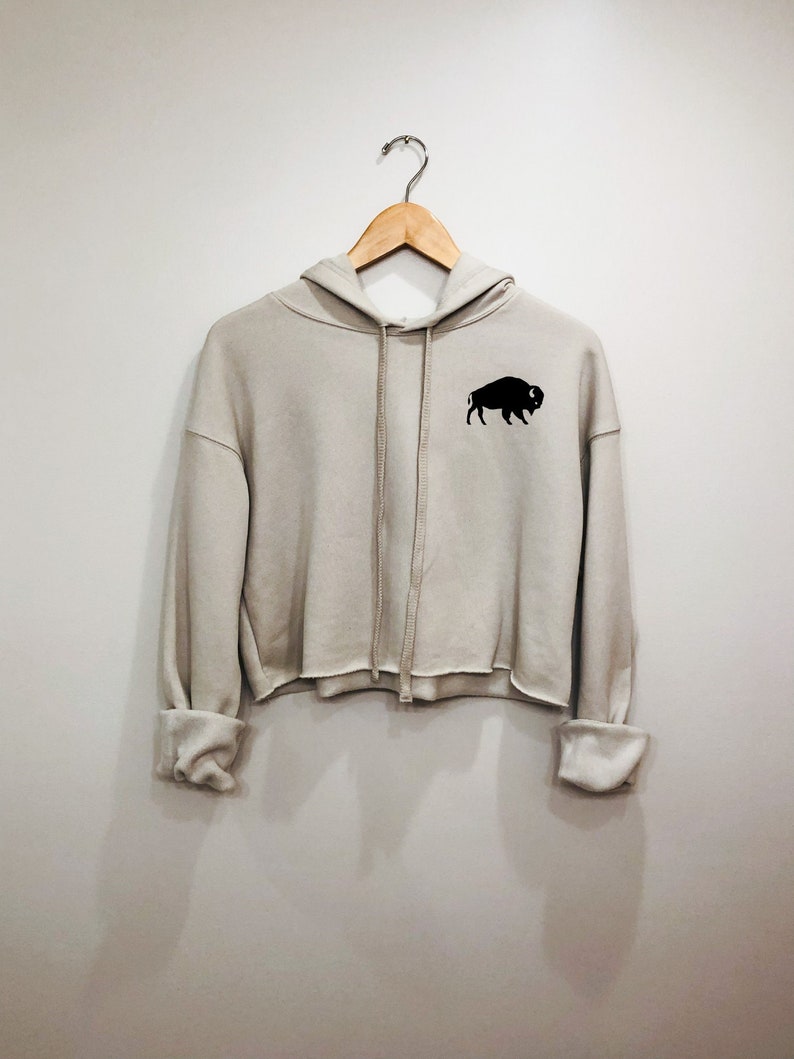 May include: A light gray cropped hoodie with a black buffalo silhouette on the left chest. The hoodie has a drawstring hood and long sleeves.