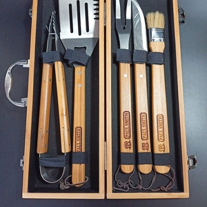Puede incluir: Un juego de herramientas para asar a la parrilla en una caja de madera con mangos de madera. Las herramientas incluyen una esp&aacute;tula, pinzas, un tenedor, un cuchillo y un pincel para untar. Los mangos est&aacute;n grabados con el texto "PAUL SMITH".