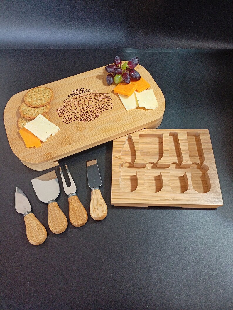 May include: A bamboo cheese board with a knife holder and five cheese knives. The board is engraved with "Diamond Anniversary 60 Years Mr. & Mrs. Roberts Est. 1963".