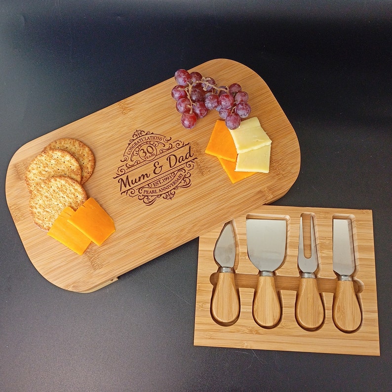 May include: A wooden cheese board with a personalised engraved message that reads "Congratulations 30 Years Mum & Dad Est. 1991 Pearl Anniversary". The board is accompanied by a set of four stainless steel cheese knives with wooden handles, all housed in a wooden case.