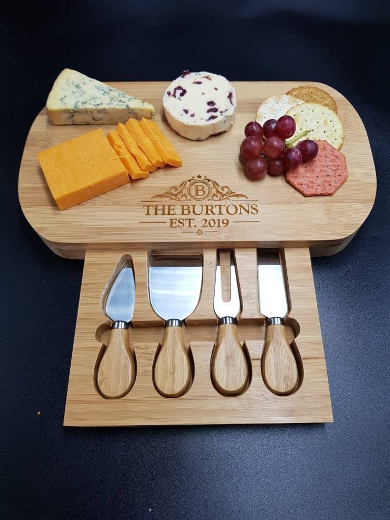 May include: A wooden cheese board with a drawer that holds four cheese knives. The board has a personalized inscription that reads "The Burtons Est. 2019". The board is filled with cheese, crackers, grapes, and a small container of cranberry sauce.