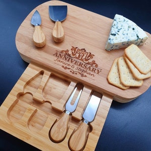 May include: A wooden cheese board with a built-in drawer for knives. The board is engraved with "50th Anniversary Janice & Stephan". The board has a wedge of blue cheese and crackers on it. There are two small knives and a cheese knife in the drawer.