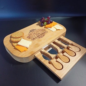 May include: A wooden cheese board with a drawer that holds four cheese knives. The board is engraved with "Diamond Anniversary 60 Years 1963 - 2023 Mr. & Mrs. Roberts". There are crackers, cheese, and grapes on the board.