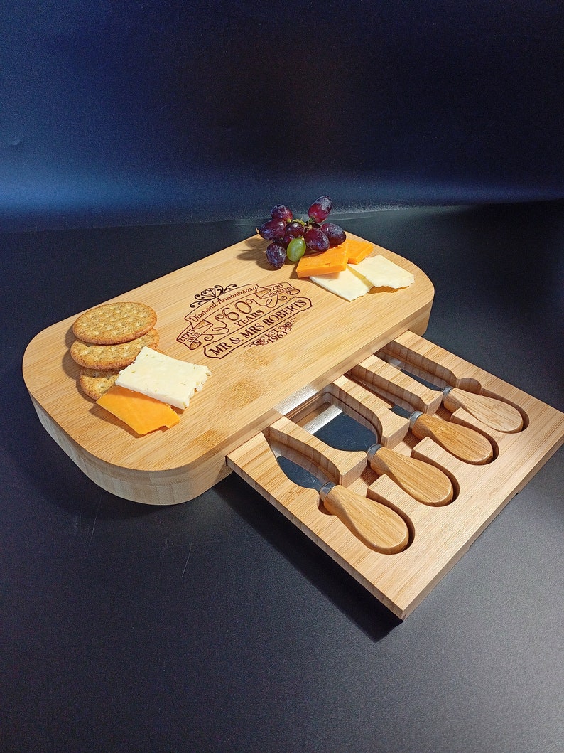 May include: A wooden cheese board with a drawer that holds four cheese knives. The board is engraved with "Diamond Anniversary 60 Years 1963 - 2023 Mr. & Mrs. Roberts". There are crackers, cheese, and grapes on the board.