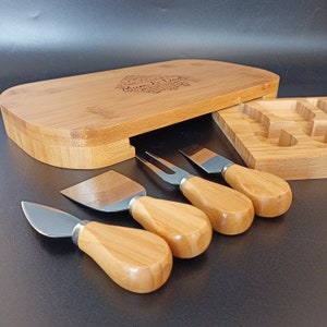 May include: A bamboo cheese board with a heart-shaped design and the text "Mum & Dad" engraved on the top. The board has a built-in drawer that holds four cheese knives with wooden handles.