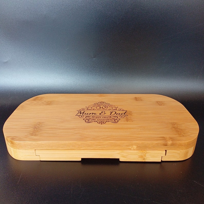 May include: A wooden chopping board with a personalised inscription that reads "Mum & Dad 30 Years Est. 1991 Pearl Anniversary". The board is made of light brown bamboo and has a rectangular shape with rounded edges.