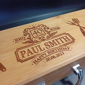 Puede incluir: Una caja de madera con una manija de metal. La caja est&aacute; grabada con el texto "BBQ King", "40 Years", "Paul Smith", "Happy Birthday" y "20.06.2023".