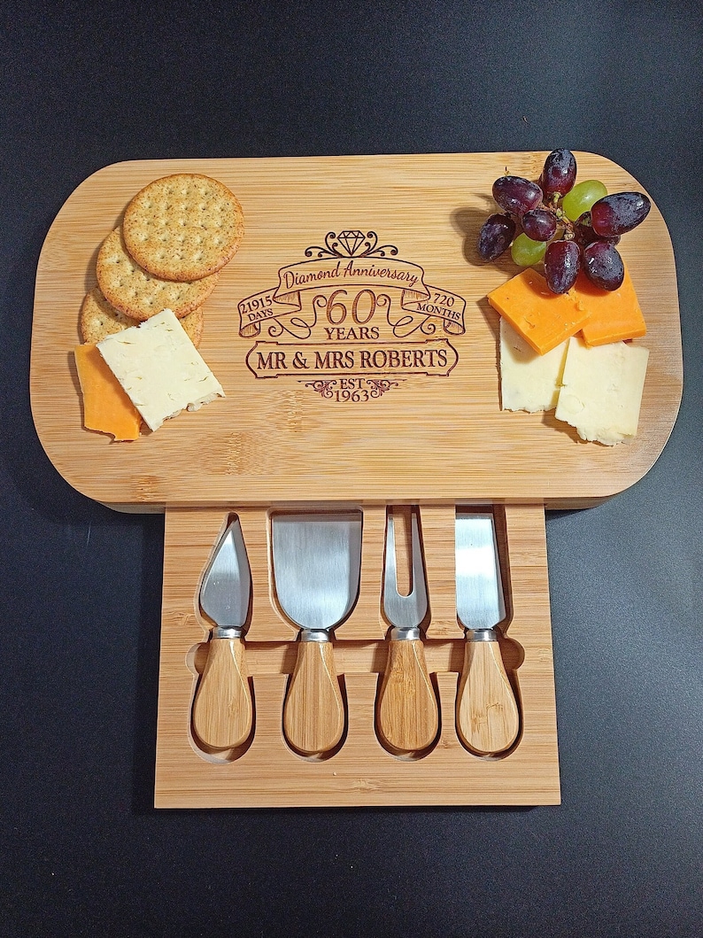 May include: A wooden cheese board with a drawer that holds four cheese knives. The board is engraved with the text "Diamond Anniversary 60 Years MR & MRS ROBERTS EST 1963". The board is also decorated with crackers, cheese, and grapes.