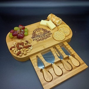 May include: A wooden cheese board with a built-in knife holder. The board is engraved with the text "July 2021 Jodie & Jordan Mr & Mrs Patterson". The board is filled with cheese, crackers, grapes, and nuts.