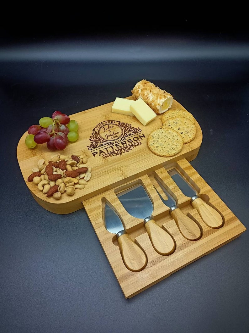 May include: A wooden cheese board with a built-in knife holder. The board is engraved with the text "July 2021 Jodie & Jordan Mr & Mrs Patterson". The board is filled with cheese, crackers, grapes, and nuts.
