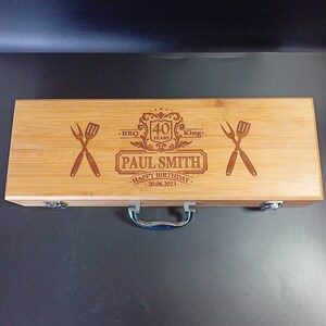 Puede incluir: Una caja de madera con asa, grabada con "BBQ King", "40 Years", "Paul Smith", "Happy Birthday" y "20.06.2023". La caja tiene dos cierres met&aacute;licos y un asa plateada.