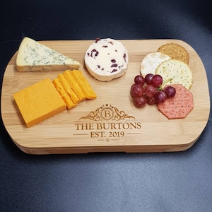 May include: A personalized bamboo cheese board with the text "The Burtons Est. 2019" engraved on the surface. The board is filled with various cheeses, crackers, grapes, and a red fruit spread.