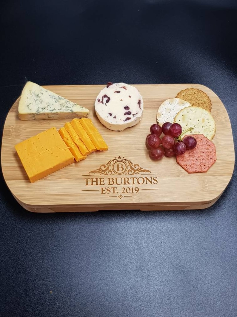 May include: A personalized bamboo cheese board with the text "The Burtons Est. 2019" engraved on the surface. The board is filled with various cheeses, crackers, grapes, and a red fruit spread.