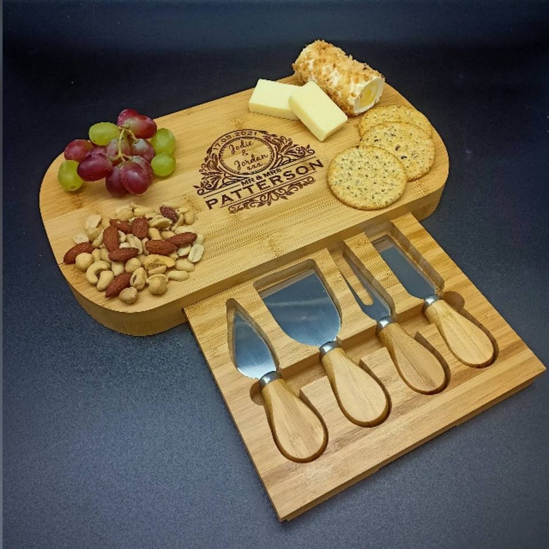 Peut inclure: Plateau &agrave; fromage en bambou personnalis&eacute; avec un tiroir coulissant contenant quatre couteaux &agrave; fromage. Le plateau est grav&eacute; du texte "July 17, 2021 Jodie & Jordan Mr. & Mrs. Patterson". Le plateau pr&eacute;sente &eacute;galement une vari&eacute;t&eacute; de collations, notamment des raisins, des noix, du fromage et des craquelins.