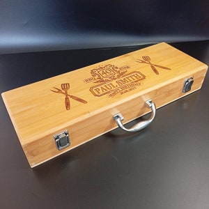 Puede incluir: Una caja de madera con una manija plateada y dos cierres. La caja est&aacute; grabada con el texto "BBQ King 40 Years Paul Smith Happy Birthday 20.06.2023".