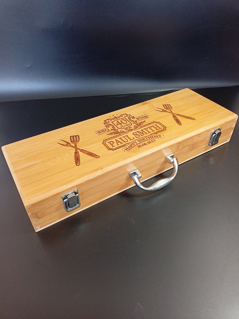 Puede incluir: Una caja de madera con una manija plateada y dos cierres. La caja est&aacute; grabada con el texto "BBQ King 40 Years Paul Smith Happy Birthday 20.06.2023".