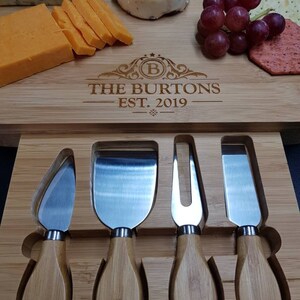 Peut inclure: Un plateau &agrave; fromage en bois avec une inscription personnalis&eacute;e "The Burtons Est. 2019" et quatre couteaux &agrave; fromage. Le plateau a une rainure pour maintenir les couteaux. Le plateau est rempli de fromage, de craquelins, de raisins et d'un petit fromage rond.