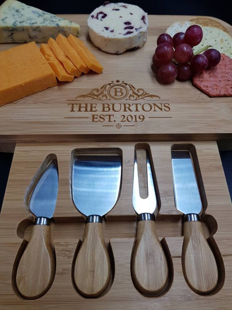 Peut inclure: Un plateau &agrave; fromage en bois avec une inscription personnalis&eacute;e "The Burtons Est. 2019" et quatre couteaux &agrave; fromage. Le plateau a une rainure pour maintenir les couteaux. Le plateau est rempli de fromage, de craquelins, de raisins et d'un petit fromage rond.
