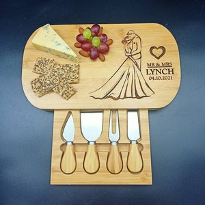 May include: A wooden cheese board with a bride and groom design, engraved with "Mr. & Mrs. Lynch 04.10.2021". The board has a built-in knife holder with four cheese knives.
