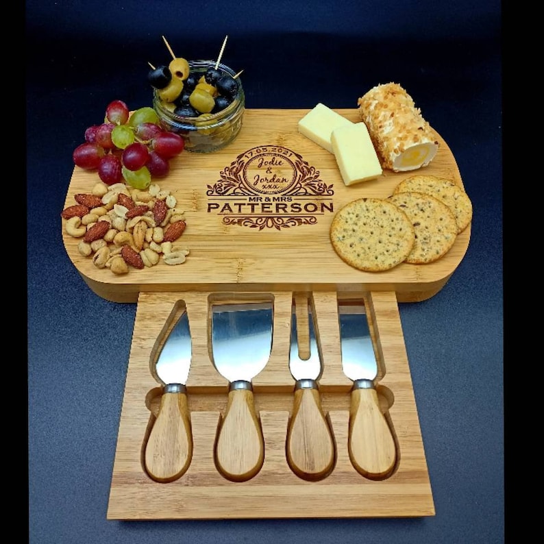 Peut inclure: Un plateau &agrave; fromage en bois avec un porte-couteaux int&eacute;gr&eacute;. Le plateau est grav&eacute; avec la date "17.05.2021", les noms "Jodie & Jordan", et le texte "Mr & Mrs Patterson". Le plateau est rempli de collations, notamment des raisins, des olives, des noix, du fromage et des craquelins.