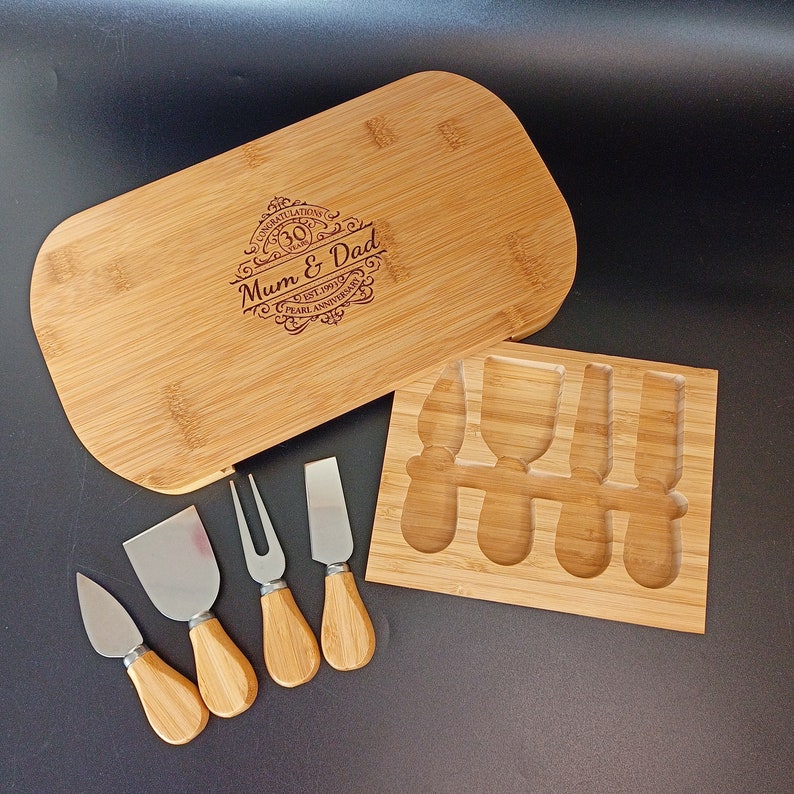 May include: A bamboo cheese board with a knife set. The board has a personalised inscription that reads "Congratulations 30 Years Mum & Dad Est. 1991 Pearl Anniversary". The knife set includes a spreader, a fork, and three knives.