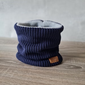 Könnte beinhalten: Ein dunkelblauer gestrickter Halsschmuck mit grauem Fleecefutter. Der Halsschmuck hat ein braunes Leder-Etikett mit dem Text "Ebbing von Bezzen"