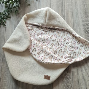 Peut inclure: Un nid pour bébé de couleur crème avec une doublure à imprimé floral. Le nid a un rabat rabattable et une petite étiquette en cuir marron avec le texte "LEBUNG COLLECTION" dessus.