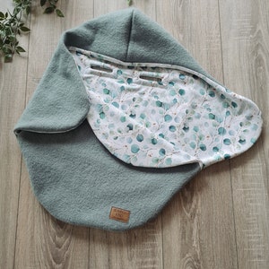 Pu&ograve; includere: Un nido per neonati blu-verde con una fodera a stampa floreale bianca e verde. Il nido ha un piccolo cartellino in pelle marrone con il testo "LEBENSO KIND" su di esso.