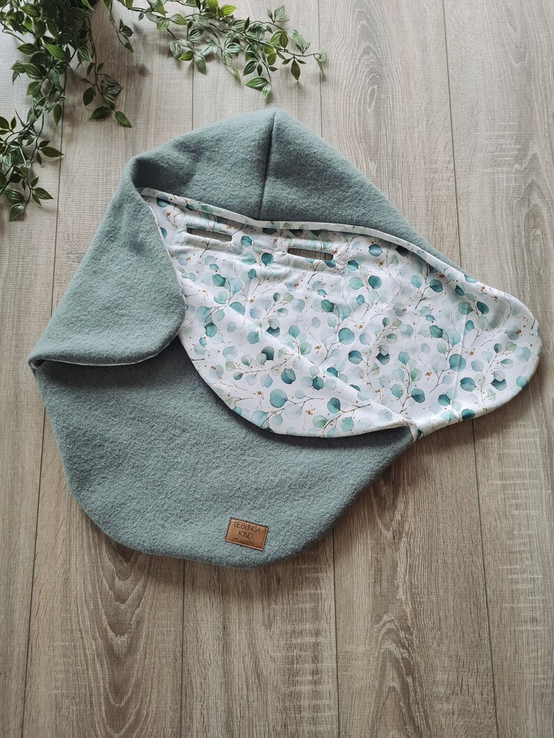 Pu&ograve; includere: Un nido per neonati blu-verde con una fodera a stampa floreale bianca e verde. Il nido ha un piccolo cartellino in pelle marrone con il testo "LEBENSO KIND" su di esso.