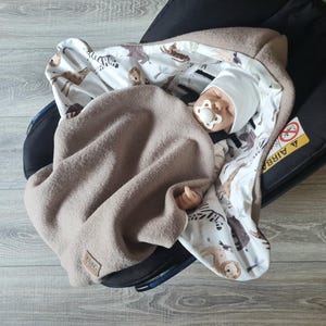 Peut inclure: Un bébé enveloppé dans une couverture en polaire taupe et une couverture à motifs avec des zèbres, des lions et d'autres animaux. Le bébé porte un bonnet blanc et a une sucette. Les couvertures sont dans un siège auto noir.
