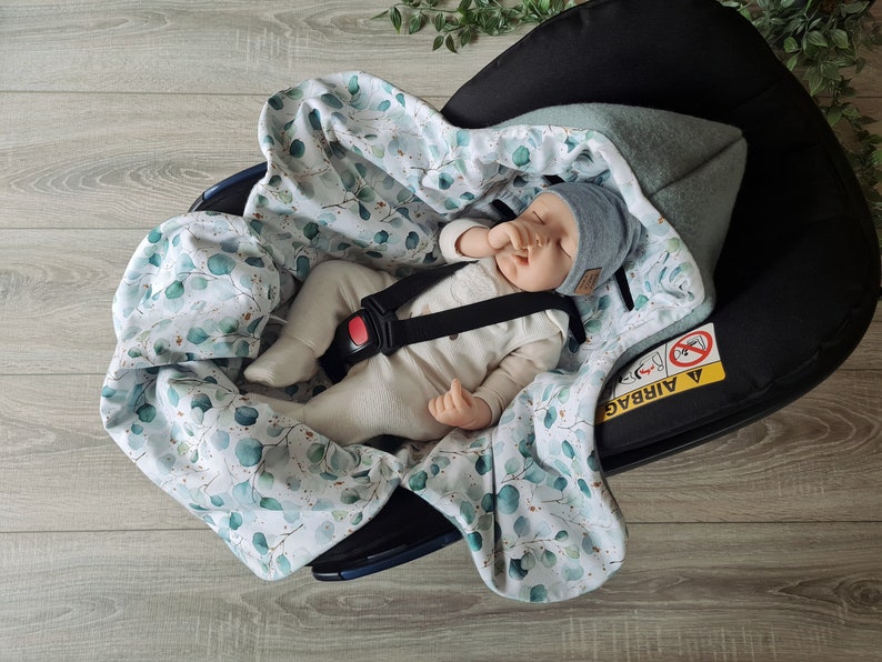 Pu&ograve; includere: Un bambino dorme in un seggiolino auto con una coperta bianca e verde con stampa di eucalipto. Il seggiolino auto ha un'etichetta di avvertenza "Airbag" nera e gialla.