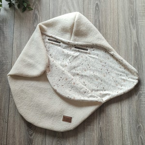 Peut inclure: Une gigoteuse blanche à capuche pour bébé avec une doublure à imprimé floral. La gigoteuse est dotée d'une étiquette en cuir marron avec le mot "NEUGUNG" imprimé dessus.
