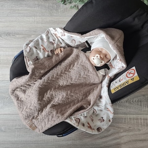 Peut inclure: Un bébé dort dans un siège auto avec une couverture matelassée marron et une couverture à capuche blanche à imprimé animalier de la forêt. Le siège auto est recouvert d'un tissu noir et porte une étiquette d'avertissement indiquant "Airbag".