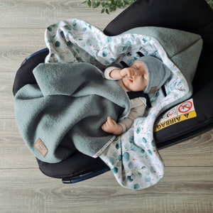 Pu&ograve; includere: Un bambino dorme in un seggiolino auto con una coperta a motivi verdi e bianchi. La coperta ha un design a foglie di eucalipto. Il seggiolino auto ha un'etichetta di avvertenza che dice "Airbag".