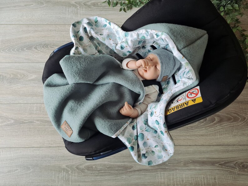 Pu&ograve; includere: Un bambino dorme in un seggiolino auto con una coperta a motivi verdi e bianchi. La coperta ha un design a foglie di eucalipto. Il seggiolino auto ha un'etichetta di avvertenza che dice "Airbag".
