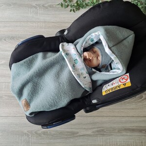 Pu&ograve; includere: Un bambino &egrave; avvolto in una morbida copertina con cappuccio verde e bianca con un motivo a foglie. Il bambino &egrave; in un seggiolino auto con la scritta "AIRBAG" visibile sul lato.