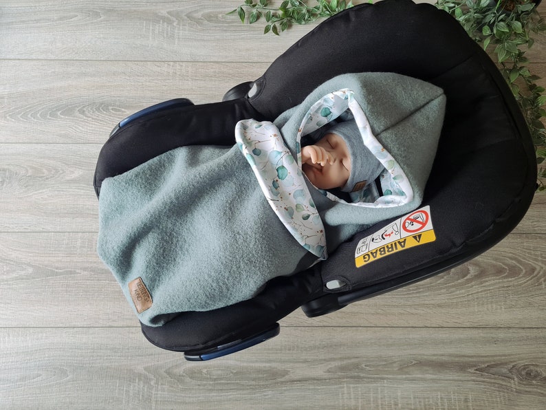 Pu&ograve; includere: Un bambino &egrave; avvolto in una morbida copertina con cappuccio verde e bianca con un motivo a foglie. Il bambino &egrave; in un seggiolino auto con la scritta "AIRBAG" visibile sul lato.