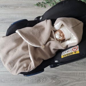 Könnte beinhalten: Ein Baby schläft in einem Autositz, eingewickelt in eine beige Babydecke mit weißem Rand. Das Baby trägt eine weiße Mütze und hat einen Schnuller im Mund. Der Autositz ist aus schwarzem Stoff und hat ein Warnschild mit der Aufschrift "AIRBAG".