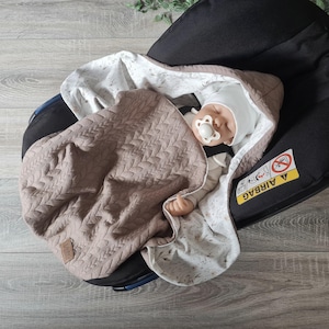 Peut inclure: Un bébé est emmailloté dans une couverture matelassée marron et blanche avec une sucette dans la bouche. Le bébé est allongé dans un siège auto noir avec le mot "AIRBAG" imprimé sur le côté.