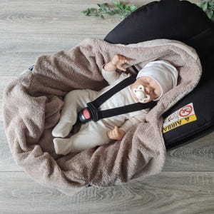 Peut inclure: Un bébé endormi dans un siège auto noir, enveloppé dans une couverture marron. Le bébé porte un bonnet et un body blancs, avec une sucette. Le siège auto est équipé d'un harnais de sécurité.